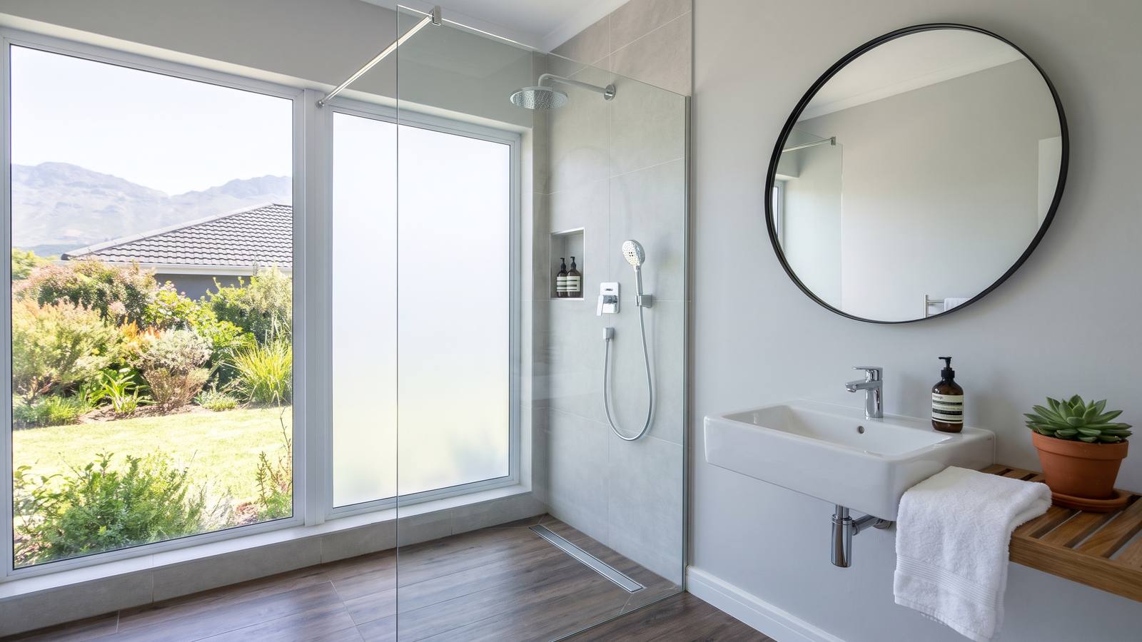 A renovated bathroom with a frameless glass walk-in shower, light grey wall tiles, a wall-hung basin and a wood-look porcelain floor in natural light.